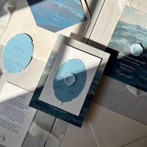 Könnte beinhalten: Elegante Hochzeitseinladungssuite mit Meeresmotiven in Blau- und Weißtönen. Die Suite umfasst Einladungskarten, RSVP-Karten und Umschläge. Die Einladungskarten haben ein Aquarell-Ozeanwellen-Design. Die Suite ist mit einem Wachssiegel versehen.