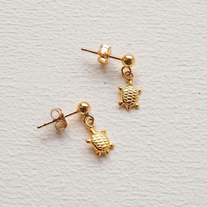 Könnte beinhalten: Ein Paar goldfarbene Ohrstecker mit einem kleinen Schildkrötenanhänger, der an jedem Ohrring baumelt. Die Ohrringe haben ein schlichtes Design.