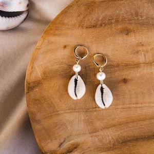 Op de afbeelding: Een paar gouden creolen met een witte schelpbedel en een witte parel die aan elke creool hangt.