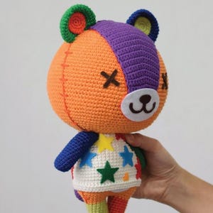 May include: A handmade crochet bear doll with an orange body, purple head stripe, and colorful accents. The bear has a white belly with star shapes and blue arms. The bear's face has a stitched X for eyes and a white nose.