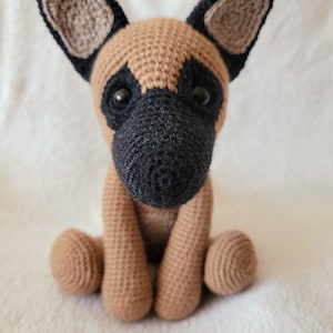 May include: A crocheted stuffed dog toy with brown and black fur. The dog is sitting with its paws tucked under its body and has a cute expression on its face.