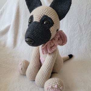 May include: A crocheted plush toy dog with beige fur, black ears, and a pink bow. The dog is sitting on a white fluffy surface.