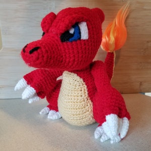 May include: A crocheted red and cream plush toy with a flame-colored tail. The toy has a fierce expression with blue eyes and white teeth.