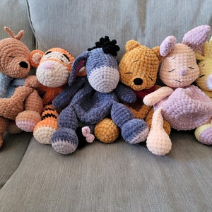 Könnte beinhalten: Eine Sammlung handgefertigter Häkeltier-Plüschtiere mit Figuren aus einer beliebten Kinderbuchreihe. Die Plüschtiere umfassen ein Känguru, einen Tiger, einen Esel, einen Bären, ein Kaninchen und ein Häschen, jeweils in ihren charakteristischen Farben und Designs.