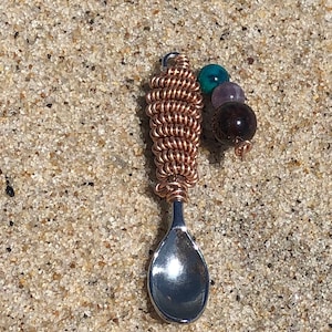 Puede incluir: Una pequeña cuchara de plata con un mango envuelto en alambre de cobre y un racimo de cuentas de colores. Las cuentas son turquesa, morado y marrón.