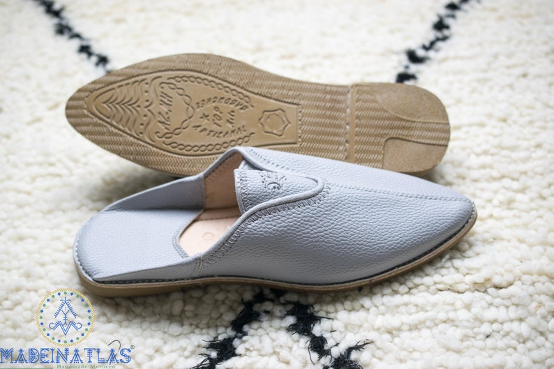 May include: A pair of gray leather slippers with a pointed toe and a brown leather sole. The sole has the text "Sandaliette" and "X For M" printed on it.