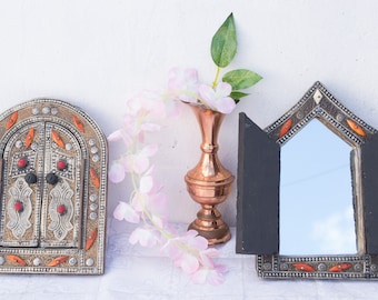 May include: Two ornate silver and orange framed mirrors with a copper vase and pink flowers in the center.