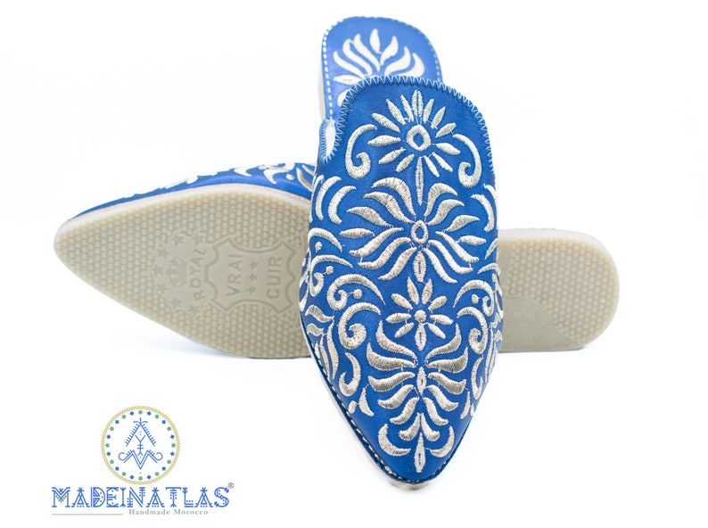 May include: A pair of blue leather slippers with silver embroidery. The slippers have a pointed toe and a flat sole. The embroidery features a floral design.