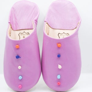 Op de afbeelding: Een paar lavendelkleurige leren slippers met kleurrijke geborduurde accenten. Elke slipper heeft een reeks kleine, decoratieve, geborduurde knopen in oranje, blauw, wit en roze. De slippers hebben een zachte, afgeronde vorm en een eenvoudig ontwerp.