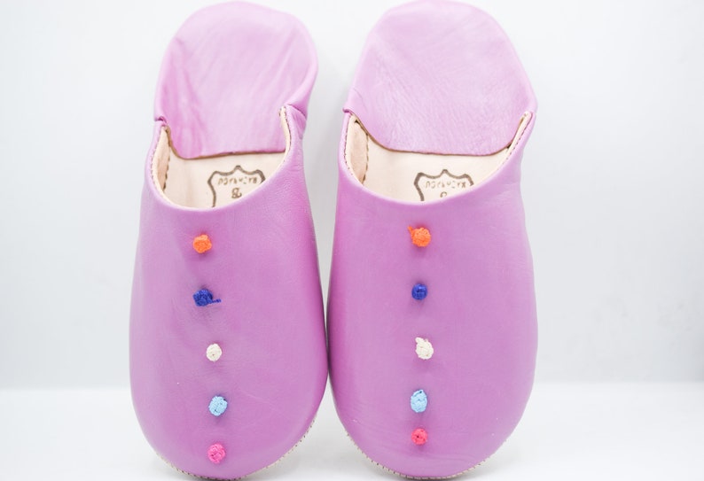 Op de afbeelding: Een paar lavendelkleurige leren slippers met kleurrijke geborduurde accenten. Elke slipper heeft een reeks kleine, decoratieve, geborduurde knopen in oranje, blauw, wit en roze. De slippers hebben een zachte, afgeronde vorm en een eenvoudig ontwerp.
