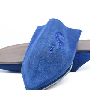 Puede incluir: Un par de zapatillas de ante azul con punta puntiaguda y una puntada decorativa en la parte superior. Las zapatillas tienen una suela de cuero marrón.