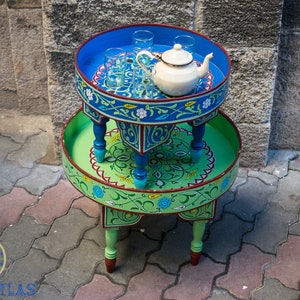 May include: A two-tiered, round, wooden table with intricate blue and green painted designs. The top tier has a white teapot and four glass cups. The table is on a gray brick surface.