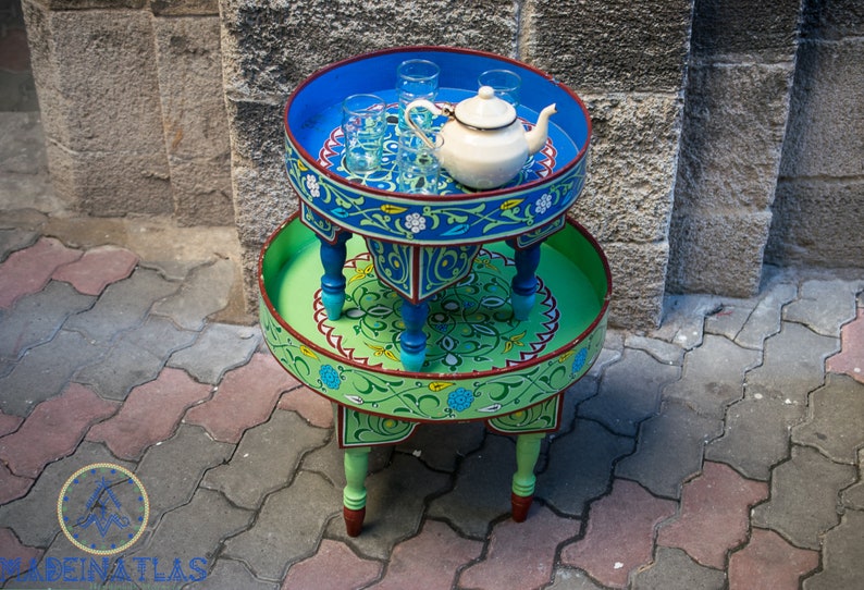 May include: A two-tiered, round, wooden table with intricate blue and green painted designs. The top tier has a white teapot and four glass cups. The table is on a gray brick surface.