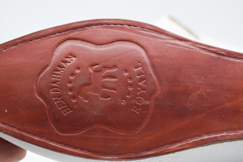 May include: Close-up of the brown leather sole of a white shoe with the text "BEN DAHMEN" and "ROYALE" embossed in a shield shape.