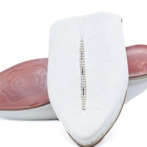 May include: White leather slippers with a pointed toe and a decorative stitch down the center. The slippers have a red leather insole.