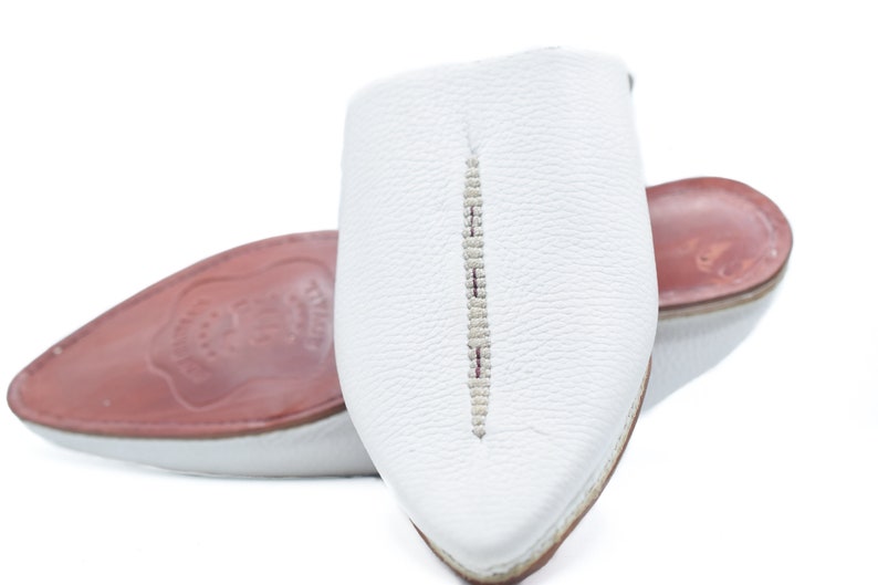 May include: White leather slippers with a pointed toe and a decorative stitch down the center. The slippers have a red leather insole.