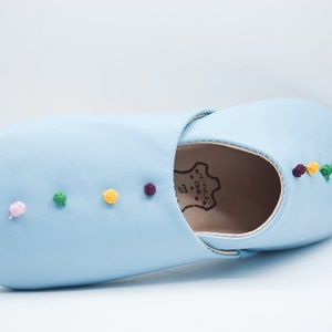 Op de afbeelding: Een lichtblauwe leren slipper met kleurrijke geborduurde accenten. De slipper heeft een ronde neus en een zachte zool. De geborduurde details omvatten kleine, ronde, kleurrijke accenten in blauw, roze, groen, geel en paars. Voor gebruik binnenshuis.