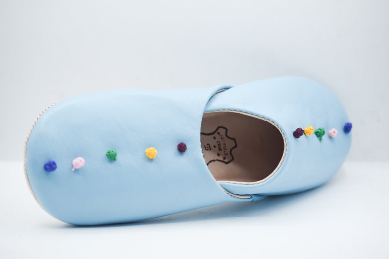 Op de afbeelding: Een lichtblauwe leren slipper met kleurrijke geborduurde accenten. De slipper heeft een ronde neus en een zachte zool. De geborduurde details omvatten kleine, ronde, kleurrijke accenten in blauw, roze, groen, geel en paars. Voor gebruik binnenshuis.