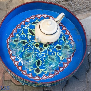 May include: A blue and white painted tray with a white teapot and five clear glasses. The tray has a decorative design with red, green, and black accents.