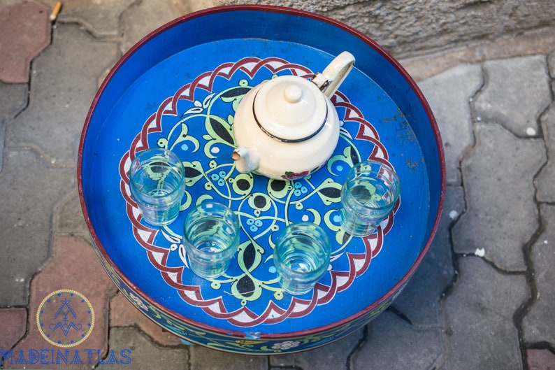 May include: A blue and white painted tray with a white teapot and five clear glasses. The tray has a decorative design with red, green, and black accents.