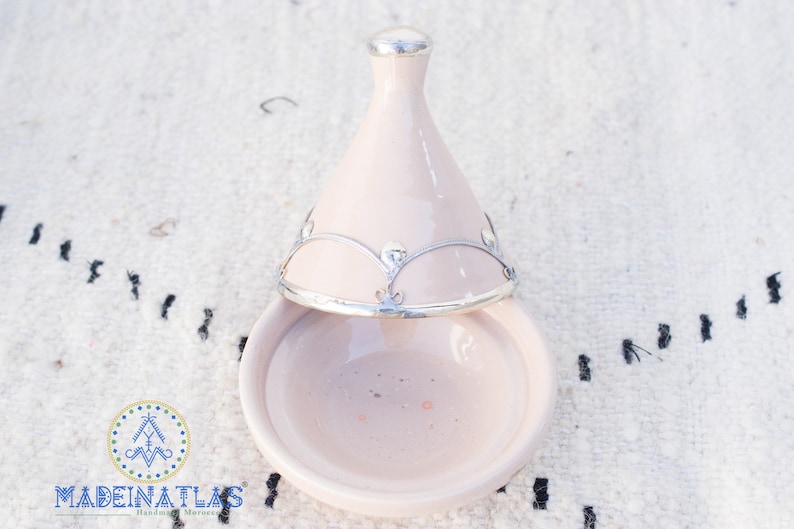 May include: A light pink ceramic tagine with a silver lid and decorative trim. The tagine is open and the lid is resting on top of the base.