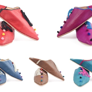 Puede incluir: Cuatro pares de zapatillas de cuero con pompones y borlas de colores. Las zapatillas son de color rojo, rosa, azul y marrón.