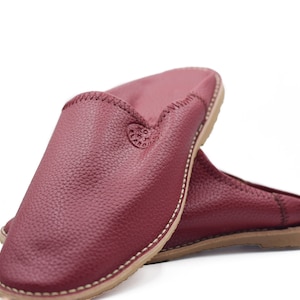 Op de afbeelding: Een paar bordeauxrode leren slippers met een gestikte naad rond de rand. De slippers hebben een gesloten neus en een platte zool.