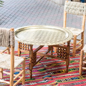 May include: A set of four wooden chairs with woven straw seats and backs, surrounding a round brass table with intricate designs. The table is set on a colourful rug with red, blue, and yellow stripes.
