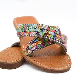 May include: A pair of brown leather sandals with a multi-colored sequined strap. The strap is criss-crossed and features a variety of colors, including red, orange, yellow, green, blue, purple, and pink.