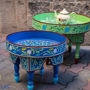 May include: Two intricately painted wooden tables with blue and green floral designs. The smaller table has a teapot and glasses on top. Both tables have a round top and four legs.