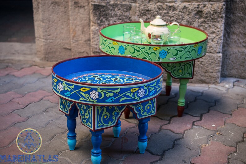 May include: Two intricately painted wooden tables with blue and green floral designs. The smaller table has a teapot and glasses on top. Both tables have a round top and four legs.