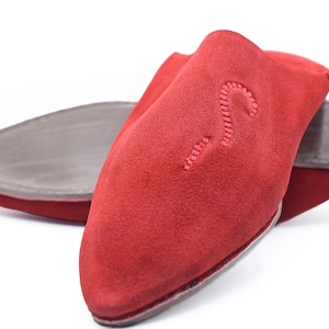 Puede incluir: Un par de zapatillas de cuero rojo con punta puntiaguda y un diseño sencillo. Las zapatillas tienen un patrón de puntadas decorativas en la parte superior.