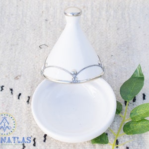May include: White ceramic tagine with a silver lid and decorative trim. The lid is open and the tagine is sitting on a white surface.