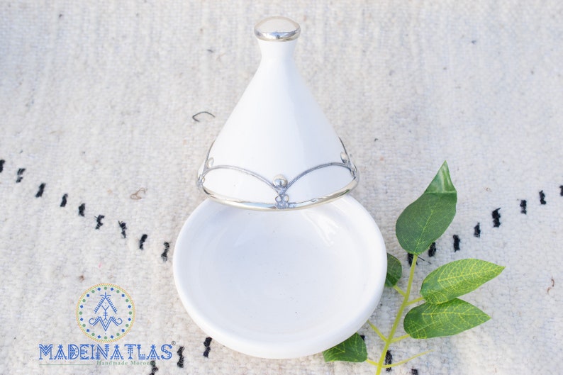 May include: White ceramic tagine with a silver lid and decorative trim. The lid is open and the tagine is sitting on a white surface.