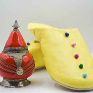 Op de afbeelding: Een rode en zilveren decoratieve container met een conische top staat naast een paar gele leren slippers. De slippers zijn versierd met kleine, kleurrijke pompons.