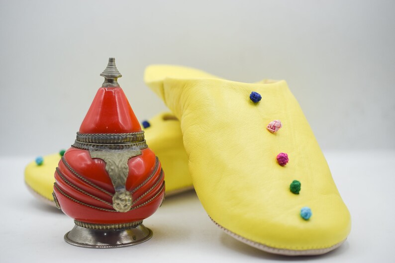 Op de afbeelding: Een rode en zilveren decoratieve container met een conische top staat naast een paar gele leren slippers. De slippers zijn versierd met kleine, kleurrijke pompons.