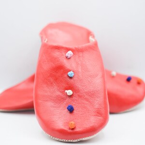 Op de afbeelding: Een paar felrode leren slippers met kleurrijke pompons. De slippers hebben een effen rode kleur met een gladde textuur en een spitse neus. De pompons zijn in verschillende kleuren, waaronder blauw, wit en oranje.