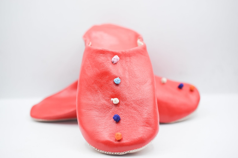 Op de afbeelding: Een paar felrode leren slippers met kleurrijke pompons. De slippers hebben een effen rode kleur met een gladde textuur en een spitse neus. De pompons zijn in verschillende kleuren, waaronder blauw, wit en oranje.