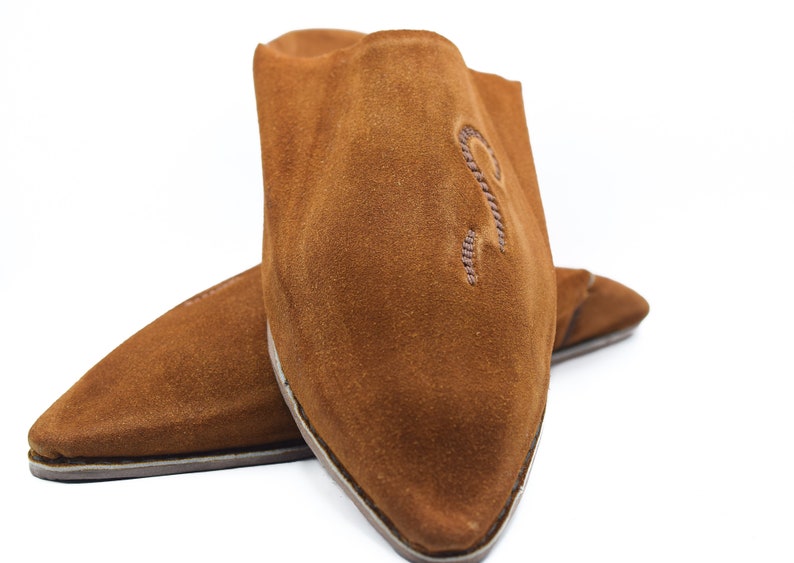 Peut inclure: Une paire de pantoufles en daim marron &agrave; bout pointu. Les pantoufles ont un design simple et sont brod&eacute;es d'une petite lettre "R" stylis&eacute;e sur le dessus.