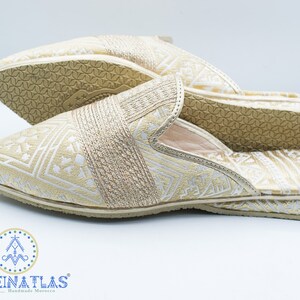 May include: A pair of gold and white patterned slippers with a closed toe and an open heel. The slippers have a geometric pattern and are made of a soft fabric.
