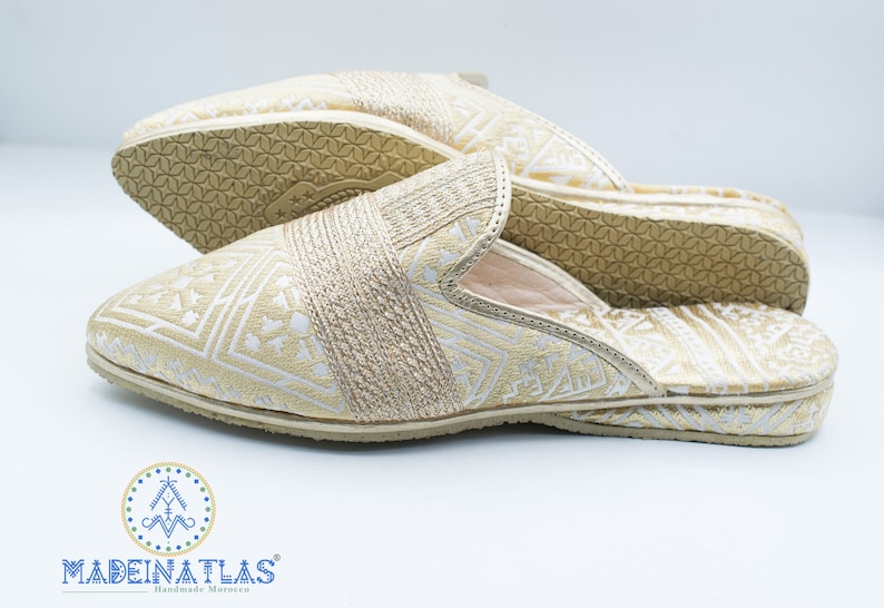 May include: A pair of gold and white patterned slippers with a closed toe and an open heel. The slippers have a geometric pattern and are made of a soft fabric.