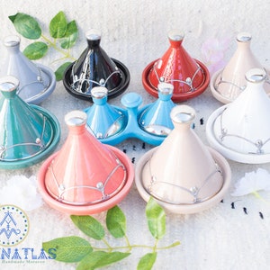 May include: A set of nine Moroccan tagines in various colors, including gray, black, red, white, teal, light blue, coral, beige, and light blue. The tagines are made of ceramic and have silver accents.