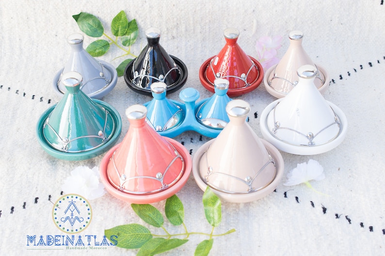 May include: A set of nine Moroccan tagines in various colors, including gray, black, red, white, teal, light blue, coral, beige, and light blue. The tagines are made of ceramic and have silver accents.