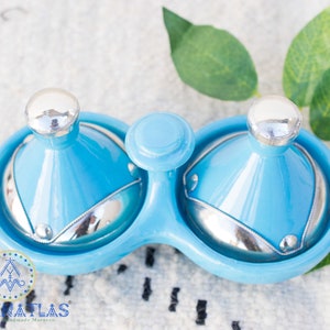 May include: A blue ceramic double dish with two lids. The lids are shaped like small tagines with silver tops. The dish is on a white surface with green leaves.
