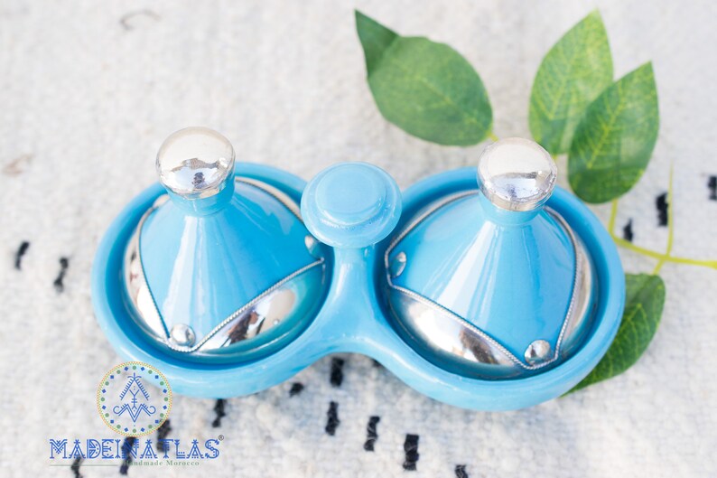 May include: A blue ceramic double dish with two lids. The lids are shaped like small tagines with silver tops. The dish is on a white surface with green leaves.