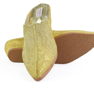 Puede incluir: Un par de zapatos dorados sin cordones con intrincados bordados. Los zapatos tienen una punta afilada y suelas marrones. El material parece brillante y texturizado. Estos zapatos probablemente están destinados a eventos especiales o ropa formal.