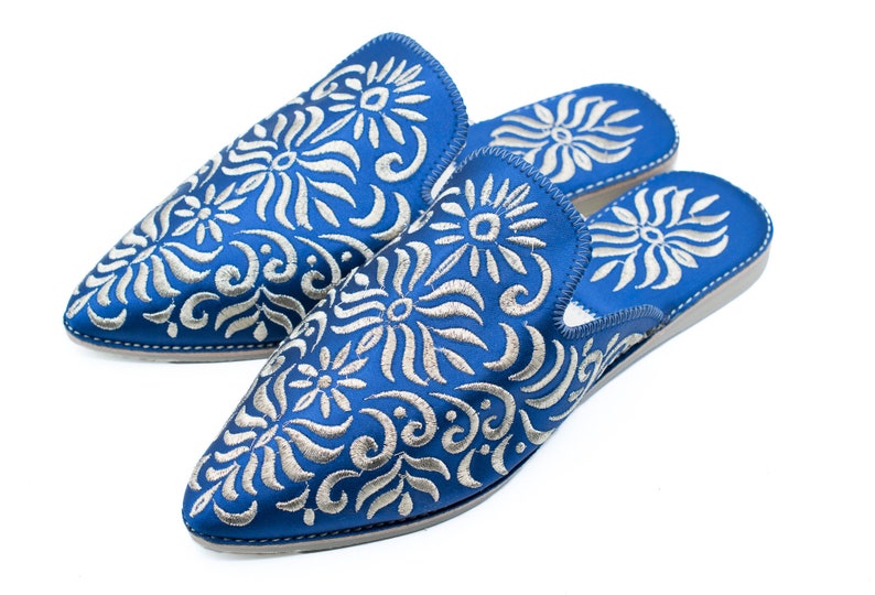 May include: A pair of blue embroidered slippers with a white floral pattern. The slippers have a pointed toe and a flat sole.