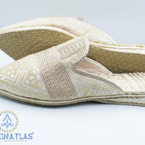 May include: A pair of gold and white patterned slippers with a closed toe and an open heel. The slippers have a geometric pattern and are made of a soft fabric. The slippers are made in Morocco.