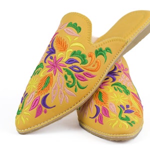 May include: Mustard yellow slip-on slippers with colorful floral embroidery. The embroidered design features pink, purple, orange, and green flowers and leaves. The slippers have a pointed toe and a soft, light-colored interior.