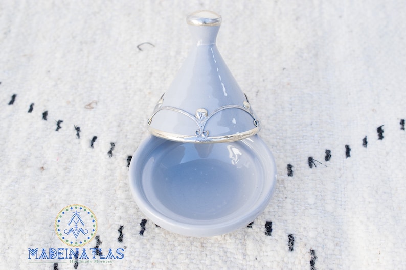 May include: A light blue ceramic dish with a lid. The lid has a silver trim and a decorative design. The dish is open and the lid is resting on top of it.